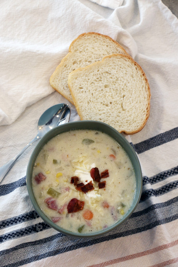 Vertical Top View Image of Ham Chowder in a Green Bowl with a spoon and two slices of bread above the bowl on a terry cloth towel.